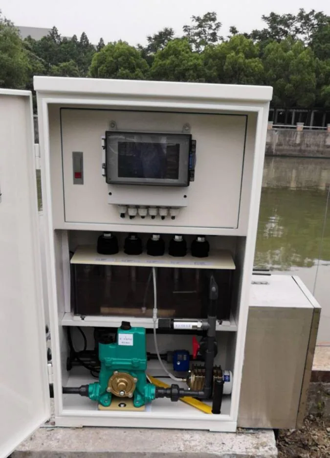 Open monitoring cabinet beside the water showing the control screen, measurement modules, sample pump, and internal piping